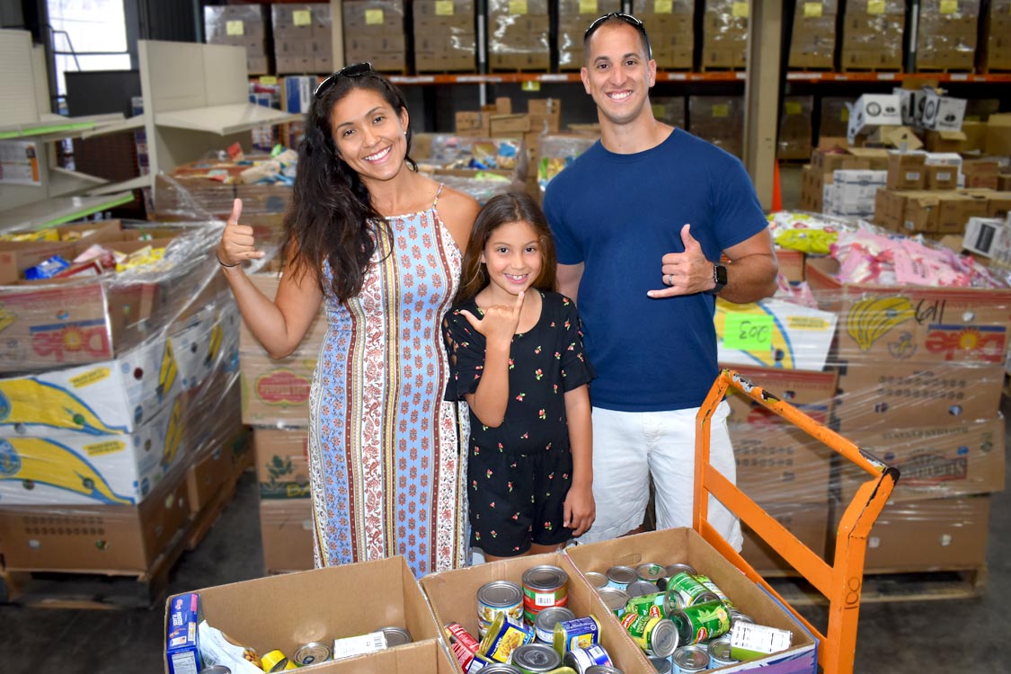 A family in Oahu happily contributing to a food drive, surrounded by donations at Hawaii Foodbank’s warehouse.