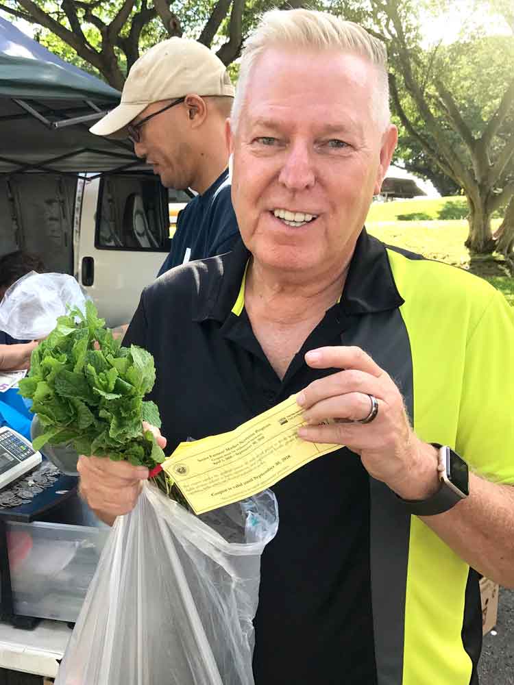 A happy participant in Hawaii's Senior Farmers Market Nutrition Program holding a bunch of fresh greens and a program coupon.